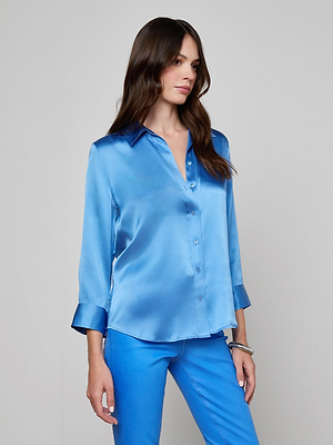 A woman with long brown hair wears a shiny regatta blue Dani Silk Blouse with long sleeves and blue pants, standing against a plain light gray background.