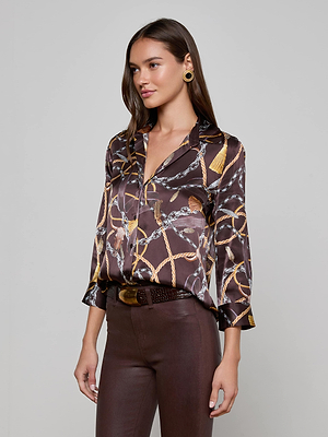 A woman with long brown hair wears the L'AGENCE Dani Silk Blouse—brown, with a Western-inspired chain print—styled with brown pants, gold hoop earrings, and a brown belt, standing against a plain light gray background.