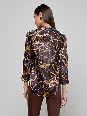 A woman with long, straight hair wears the L'AGENCE Dani Silk Blouse in dark brown, featuring a Western chain print with gold and silver details. She pairs it with dark brown pants and gold earrings, facing away from the camera.