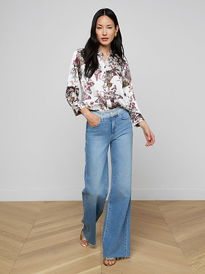 A woman with long black hair stands indoors, confidently posing on a light wooden floor against a white wall. She wears the Dani Silk Blouse with a butterfly jungle print and light blue wide-leg jeans, with one hand in her pocket.