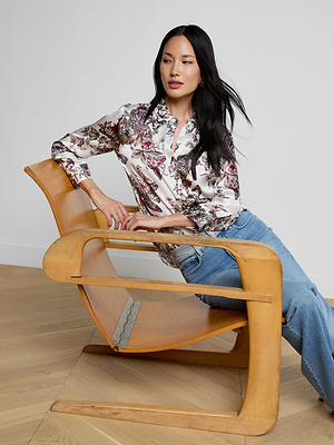 A woman with long dark hair, wearing a Dani Silk Blouse in butterfly jungle print and blue jeans, sits sideways on a modern wooden chair in a room with light wooden floors and a white wall.