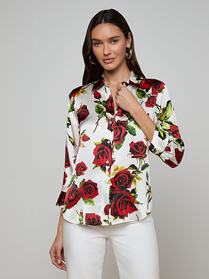 A woman with long brown hair wears a Dani Silk Blouse with a red rose print and white pants, standing against a plain light gray background. She smiles softly while gently touching her collar.