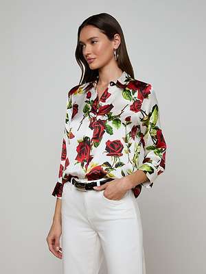 A woman models the Dani Silk Blouse in a red rose print, paired with white pants and a black belt, standing before a plain light gray background.
