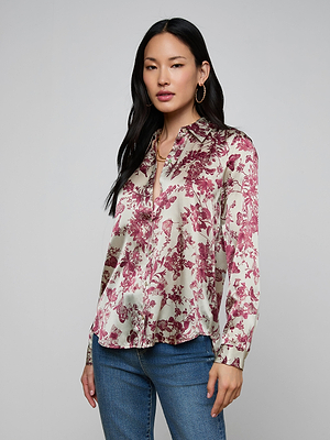 A woman with long black hair wears the Tyler Silk Blouse, a cream button-down with a burgundy floral butterfly print, and blue jeans. She stands against a plain gray background, looking at the camera with a neutral expression.