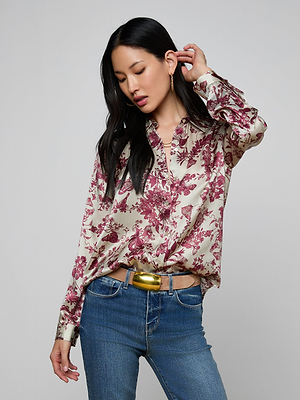 A woman with long dark hair wears the Tyler Silk Blouse, featuring a red floral butterfly print. Paired with blue jeans and a gold belt, she poses touching her hair and looks down against a plain light background.