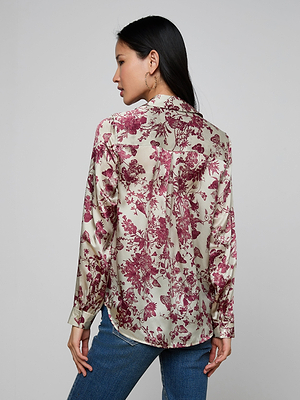 A woman with long dark hair wears the Tyler Silk Blouse, a button-down featuring a red floral butterfly print. With her back turned and head slightly to the side, she pairs it with blue jeans against a light gray background.
