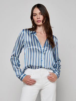 A woman with long brown hair wears the L'AGENCE Tyler Silk Blouse in a blue stripe print and white pants, standing against a plain light gray background with one hand in her pocket.