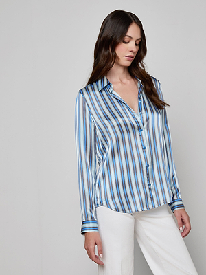 A woman with long brown hair wears the Tyler Silk Blouse, a shiny blue stripe-print button-down by L'AGENCE, paired with white pants against a plain light gray background.