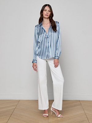 A woman with long brown hair and a neutral expression stands indoors against a plain light wall, wearing the Tyler Silk Blouse in blue stripe print, white wide-leg pants, and white heels.