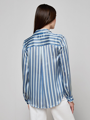 A woman with long brown hair is seen from behind wearing the Tyler Silk Blouse, a shiny L’AGENCE button-down with vertical blue stripes, paired with white pants against a plain light background.