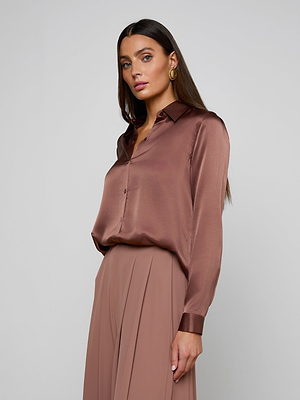 A woman with long dark hair wears a shiny, brown Tyler Silk Blouse and high-waisted, pleated taupe pants, standing against a plain light gray background and looking at the camera with a neutral expression.