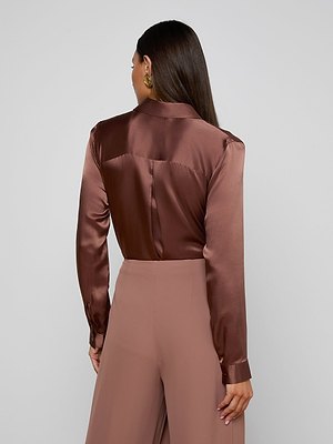 A woman with long dark hair, seen from behind, wears the shiny brown Tyler Silk Blouse by L’AGENCE with long sleeves and high-waisted, wide-legged taupe pants, standing against a plain light background.