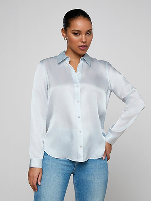 A woman wearing the Tyler Silk Blouse in light blue and blue jeans stands against a plain light background, one hand on her hip, looking confidently at the camera.