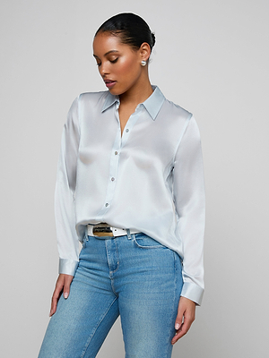 A woman in a light blue Tyler Silk Blouse and blue jeans stands against a plain background, looking down with her hand on her hip.