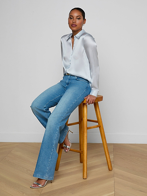 A woman sits on a wooden stool, wearing the light blue Tyler Silk Blouse, blue jeans, and silver high-heeled sandals. She poses confidently against a plain white wall and wooden floor.