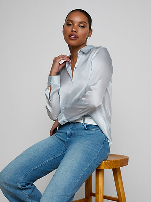A woman in a light blue Tyler Silk Blouse and blue jeans poses confidently on a wooden stool, one hand on her leg and the other touching her neck, set against a plain light gray background.