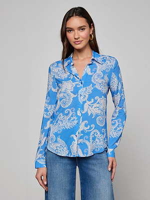 A woman smiles gently at the camera, wearing the Holly Blouse in regatta blue with a white paisley print and wide-leg blue jeans, standing against a plain light gray background.