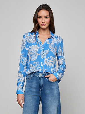 A woman with long brown hair wears the Holly Blouse in regatta blue paisley print, tucked into high-waisted jeans. She stands against a plain light gray background, looking at the camera with a slight smile.