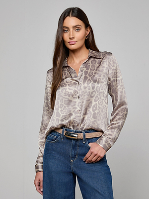 A woman with long brown hair wears the silky Tyler Silk Blouse in gray leopard print and blue jeans, standing against a plain gray background and looking confidently at the camera with one hand in her pocket.