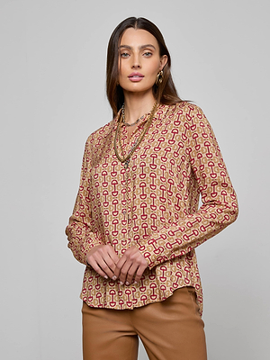 A woman in the L'AGENCE Holly Blouse—a slim fit, long sleeve button-down with red and beige designs—wears layered necklaces and tan pants as she stands against a plain gray background, gazing slightly to the side.