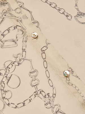 Close-up of the Tyler Silk Blouse, a beige button-down by L'AGENCE, featuring a chain print with jewelry outlines and three small white buttons along the seam.