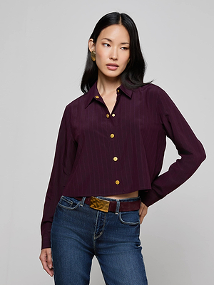 A woman with long dark hair wears the Cosette Pinstriped Shirt in maroon, paired with blue jeans, a brown belt, and gold earrings. She stands with one hand on her hip, gazing to the side.