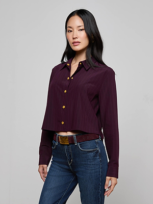 A woman stands against a plain background wearing the Cosette Pinstriped Shirt, a tailored dark purple button-up with gold buttons, paired with blue jeans and a brown belt. Her straight black hair is worn down as she looks confidently at the camera.