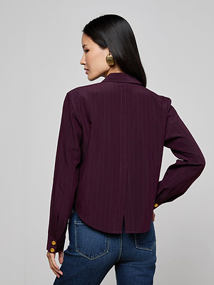 A woman with long black hair wears the Cosette Pinstriped Shirt with gold buttoned cuffs and blue jeans, standing facing away and glancing to the side against a plain background.
