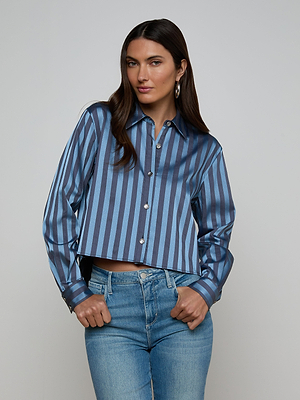 A woman with long dark hair wears the Cosette Satin Shirt, featuring blue stripes and a high-low hem, paired with light blue jeans. She stands against a plain light gray background, one hand in her pocket and a relaxed expression.