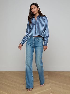 A woman with long brown hair stands indoors on a wooden floor, looking confidently at the camera. She wears the Cosette Satin Shirt—a blue striped cropped top with a high-low hem—paired with light blue high-waisted jeans and open-toe sandals.