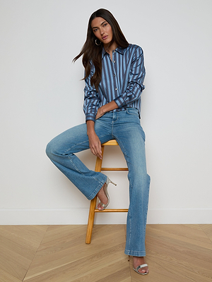 A woman with long brown hair sits on a wooden stool against a white wall, wearing the Cosette Satin Shirt, light blue flared jeans, and silver open-toe heels. She poses with a relaxed and confident demeanor.