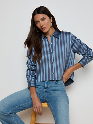 A woman with long brown hair sits on a wooden stool against a white background, wearing the Cosette Satin Shirt—blue with stripes, featuring a high-low hem—paired with light blue jeans.