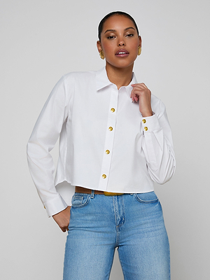 A woman stands confidently against a light gray background, wearing the Cosette Shirt—an organic cotton poplin blouse with gold buttons—and blue jeans, her hand resting near the collar as she looks at the camera.