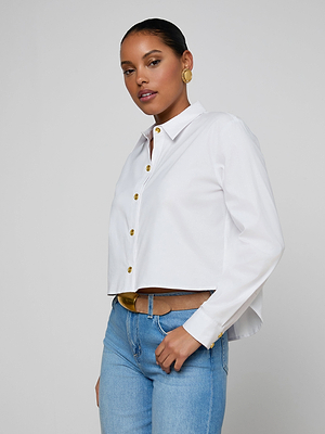 A woman wearing the white Cosette Shirt with gold buttons, paired with blue jeans and gold earrings, stands against a plain gray background, gazing confidently at the camera.