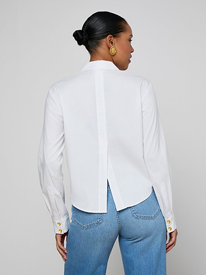 A woman with her hair in a low bun wears the Cosette Shirt—white organic cotton poplin with gold cuff buttons—paired with blue jeans. She faces away, highlighting the shirt’s central pleat and curved hem.