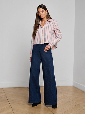 A woman with long brown hair and a neutral expression stands indoors on a wooden floor, wearing the Cosette Shirt—an organic cotton-blend pink and white striped tailored shirt—and high-waisted, wide-leg dark blue jeans with black shoes.