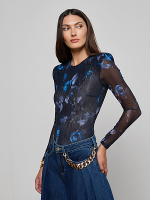 A woman with long brown hair models the Trista Bodysuit, a stretch mesh midnight floral design with structured shoulder pads, paired with high-waisted blue jeans and a gold chain belt. She stands against a plain light gray background, looking to the side.