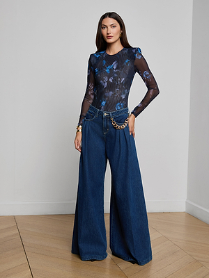 A woman with long brown hair and structured shoulder pads stands indoors wearing the Trista Bodysuit—a midnight floral, stretch mesh piece—paired with high-waisted, wide-leg blue jeans, a gold chain belt, and a neutral expression.