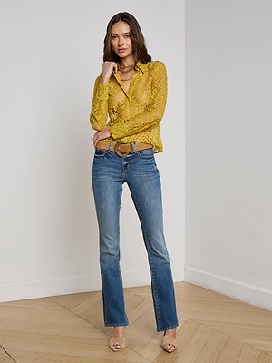 A woman stands indoors on a wood floor, wearing the Maia Floral Lace Blouse by L’AGENCE in mustard yellow, paired with blue jeans, a tan belt, beige heeled sandals, layered necklaces, and her long brown hair down.