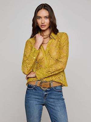 A woman with long brown hair wears the L'AGENCE Maia Floral Lace Blouse in mustard yellow, paired with blue jeans and gold necklaces, standing against a plain light gray background.