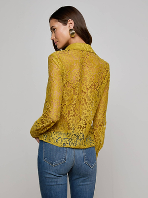A woman faces away, wearing the L'AGENCE Maia Floral Lace Blouse in mustard yellow with long sleeves and blue jeans. Her dark hair is tucked behind her ear, complemented by large gold hoop earrings.