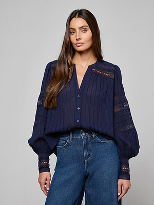 A woman with long brown hair models the Kiera Blouse, a navy blue bohemian top with lace details, paired with blue jeans. She stands against a plain gray background, looking directly at the camera with a neutral expression.