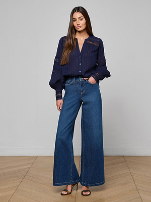 A woman with long brown hair models the Kiera Blouse, a navy blue bohemian button-up with long sleeves, paired with wide-leg blue jeans and black heeled sandals. She stands on a wooden floor against a light gray background.