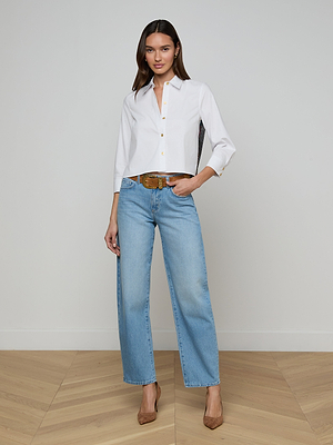 A woman stands in a minimal room wearing the Fawn Floral Print Shirt, light blue wide-leg jeans, a brown belt, and tan high heels—one hand on her hip and a neutral expression.