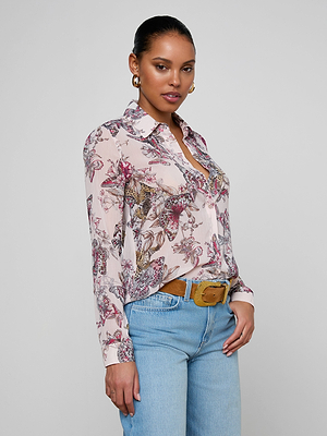 A woman stands against a plain background, confidently wearing the Laurent Blouse—a relaxed fit, long sleeve butterfly jungle print sheer poly chiffon top—paired with light blue jeans, a brown belt, and gold hoop earrings. Her hair is pulled back.