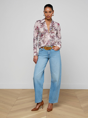 A woman poses confidently in the Laurent Blouse, a sheer light pink floral poly chiffon top with relaxed long sleeves, paired with light blue high-waisted jeans, a brown belt, and brown heeled shoes. One hand rests in her pocket.
