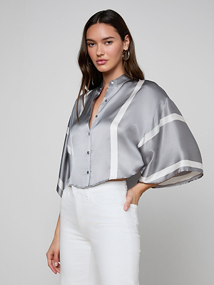 A woman with long brown hair wears the Dru Silk Blouse— a loose, silky gray top with kimono sleeves— and white high-waisted pants, standing sideways and looking at the camera against a plain gray background.