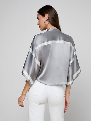 A woman with long brown hair, seen from behind, wears the Dru Silk Blouse—a loose, shiny silver silk top with white sleeve and back stripes—paired with high-waisted white pants.