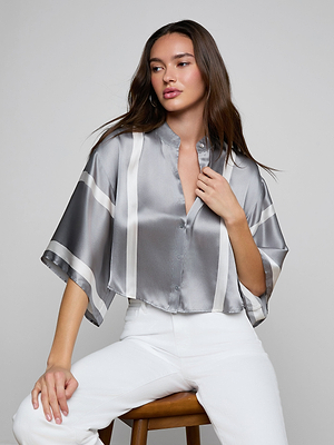 A woman with long brown hair and a neutral expression sits on a wooden stool against a light gray background, wearing the Dru Silk Blouse—a shiny, short-sleeved blouse with silver stripes—and white pants.