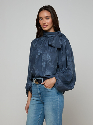 A woman with long dark hair wears the Rhett Oversized Blouse—blue jacquard with a neck bow and voluminous sleeves—paired with light blue jeans and a black belt, standing against a plain gray background.